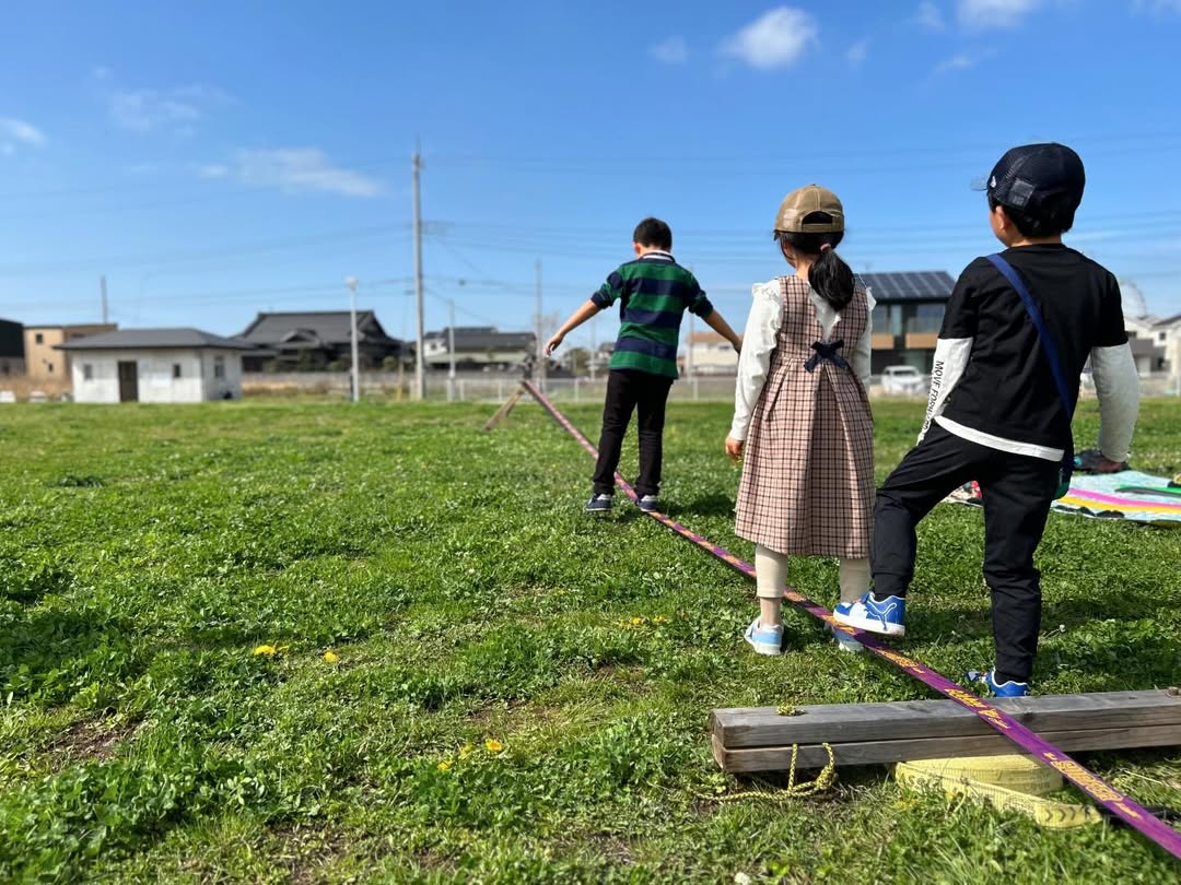 青空の下、芝生に張ったスラックラインを順番に歩く子どもたち